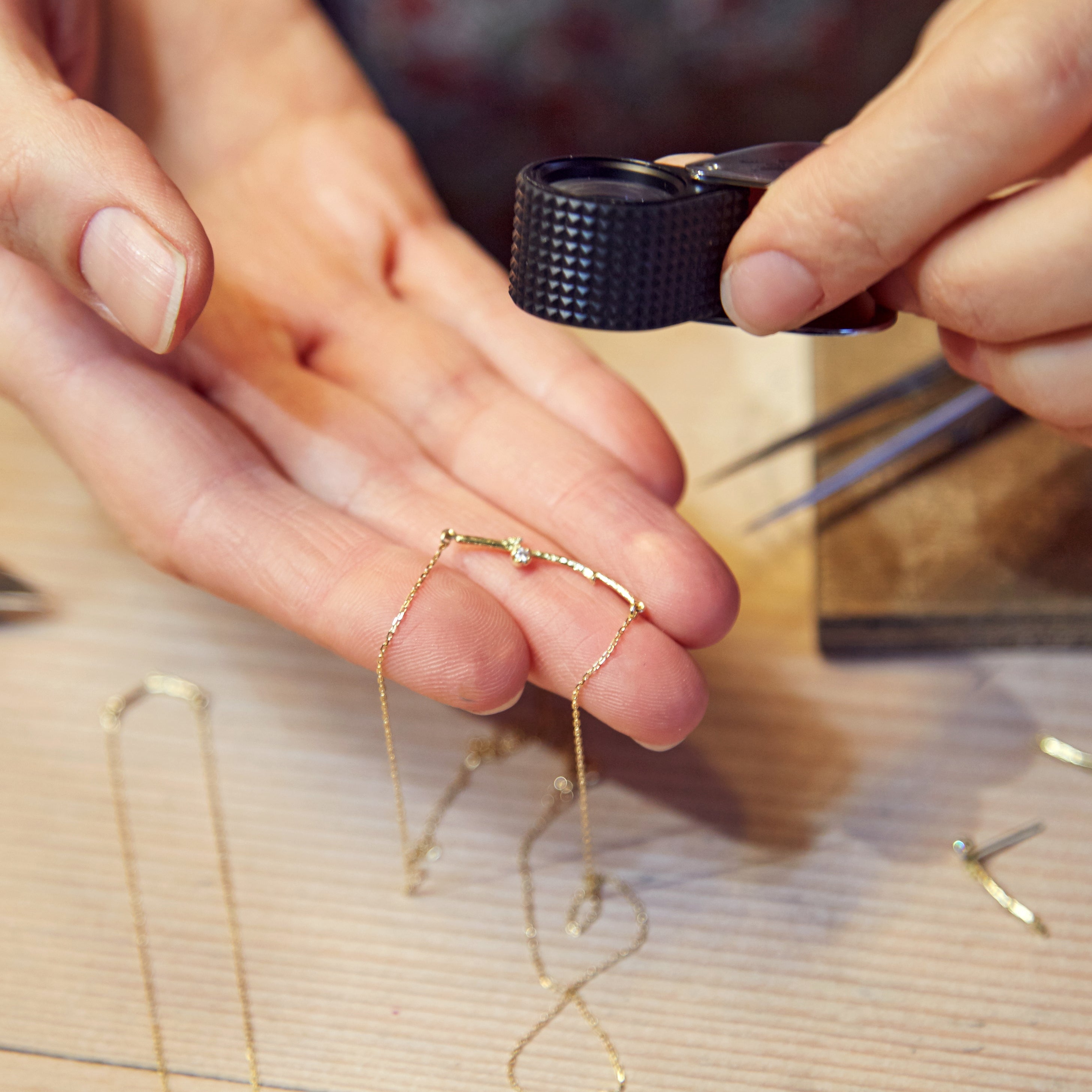 Close-up of a person's hands holding a delicate gold chain necklace while inspecting it with a black magnifying loupe on a wooden workbench.