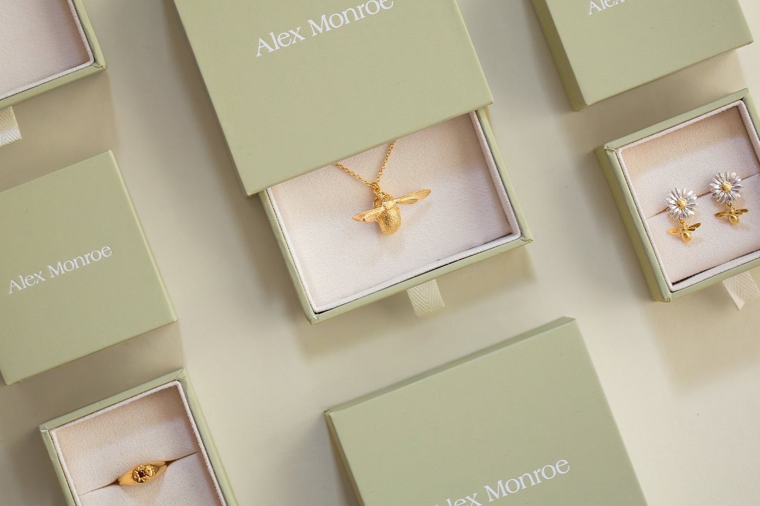 An overhead flat-lay of Alex Monroe jewelry packaging and pieces, showing a gold bee pendant necklace, a pair of gold and white flower stud earrings, and a gold ring nestled in light sage green presentation boxes.