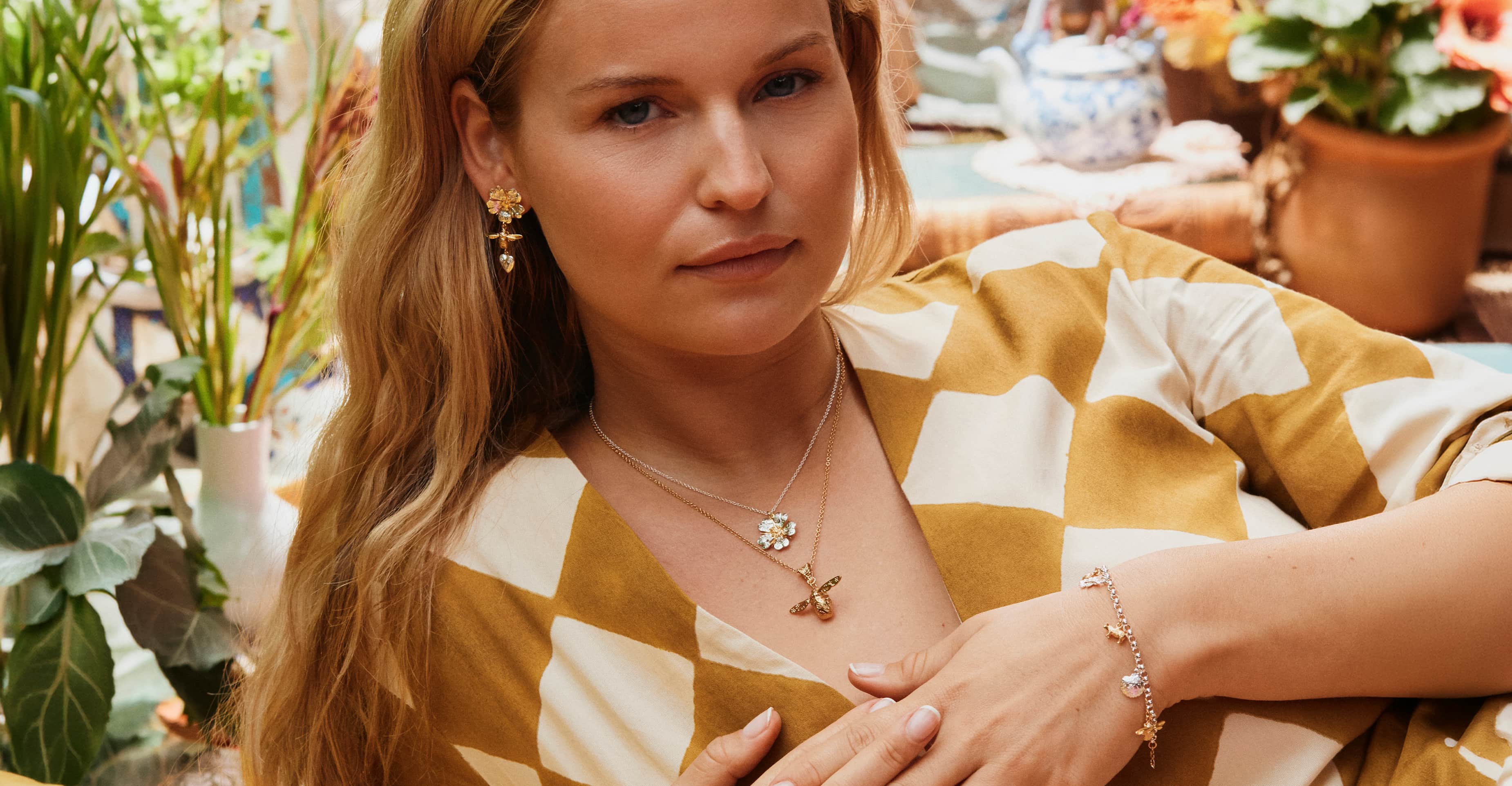 Woman in a patterned dress wearing gold flower earrings, layered necklaces and a delicate charm bracelet, surrounded by plants