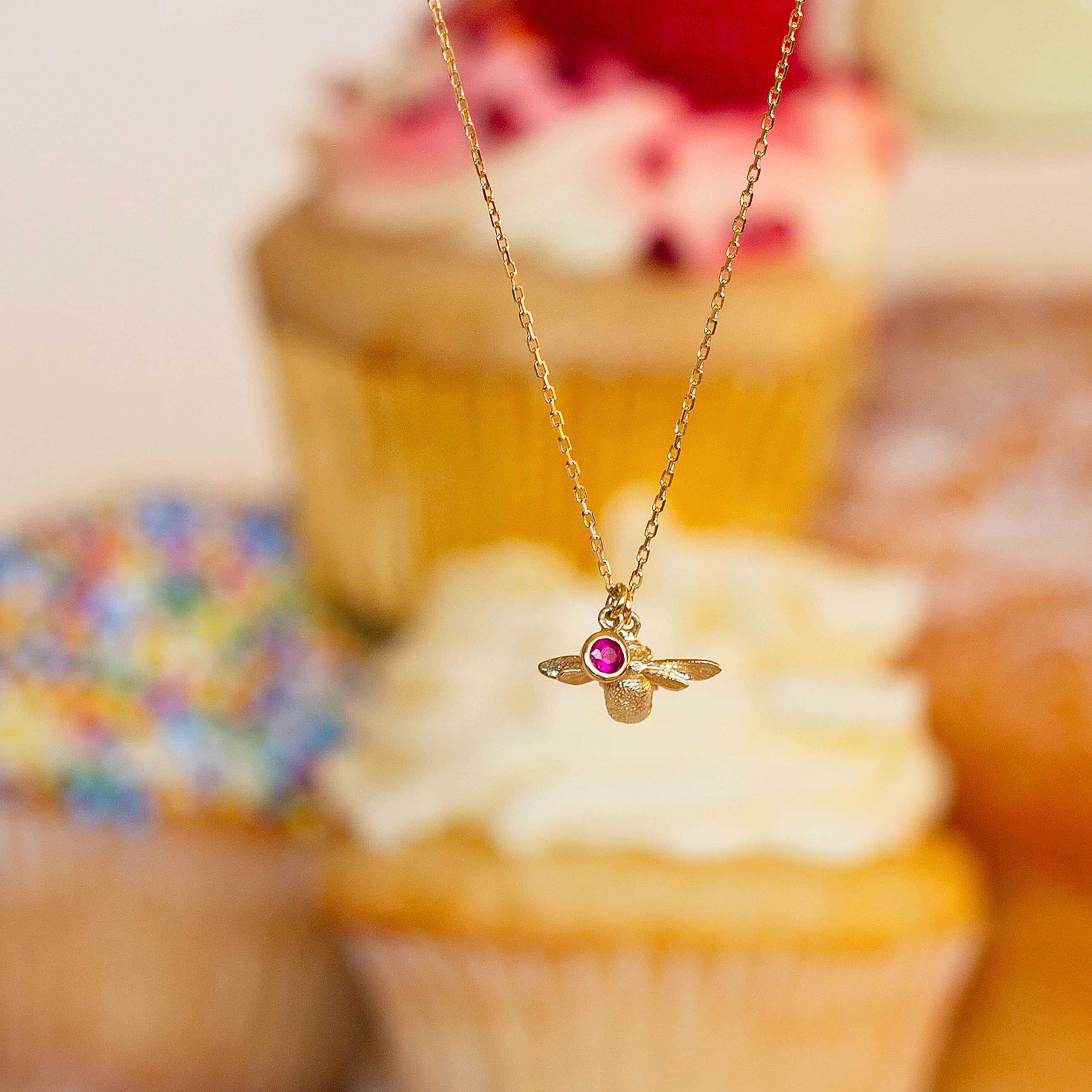 Alex Monroe ruby bee pendant necklace in 18ct gold displayed in front of cupcakes to celebrate the 40th anniversary collection