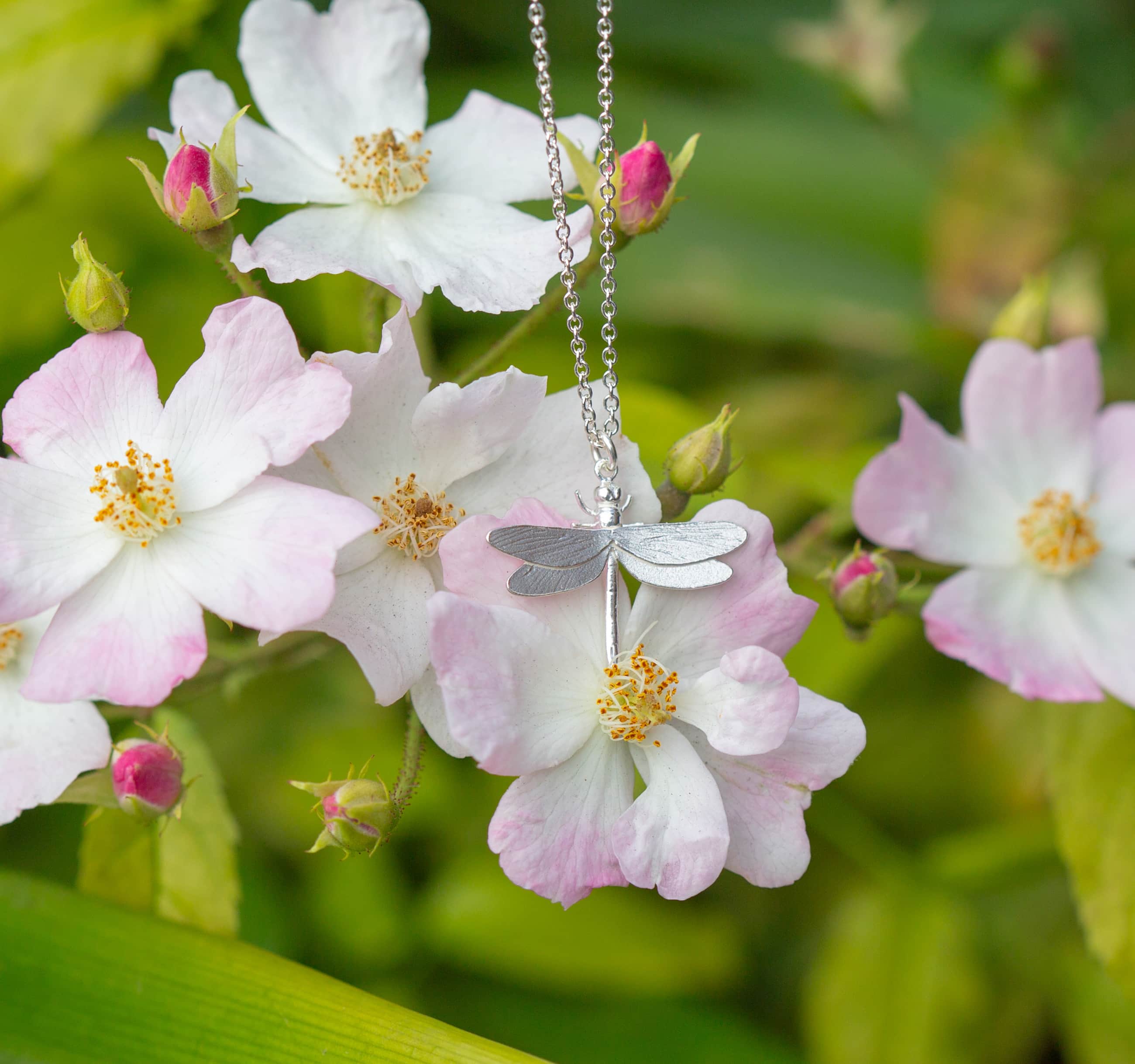 A finely detailed dragonfly pendant rests among soft blossoms and fresh greenery, capturing a quiet moment in nature