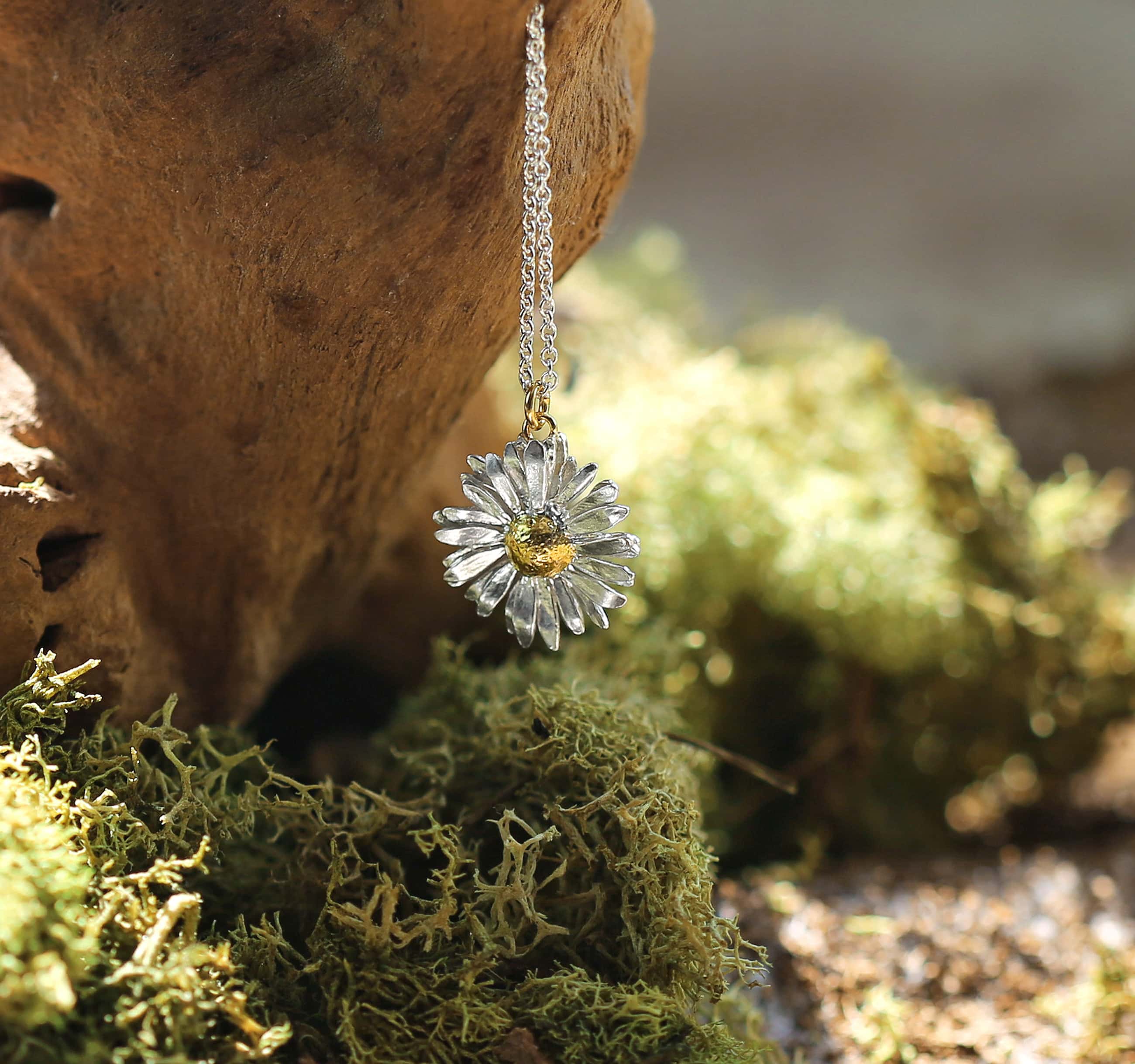 Silver daisy flower pendant necklace hanging on chain against moss