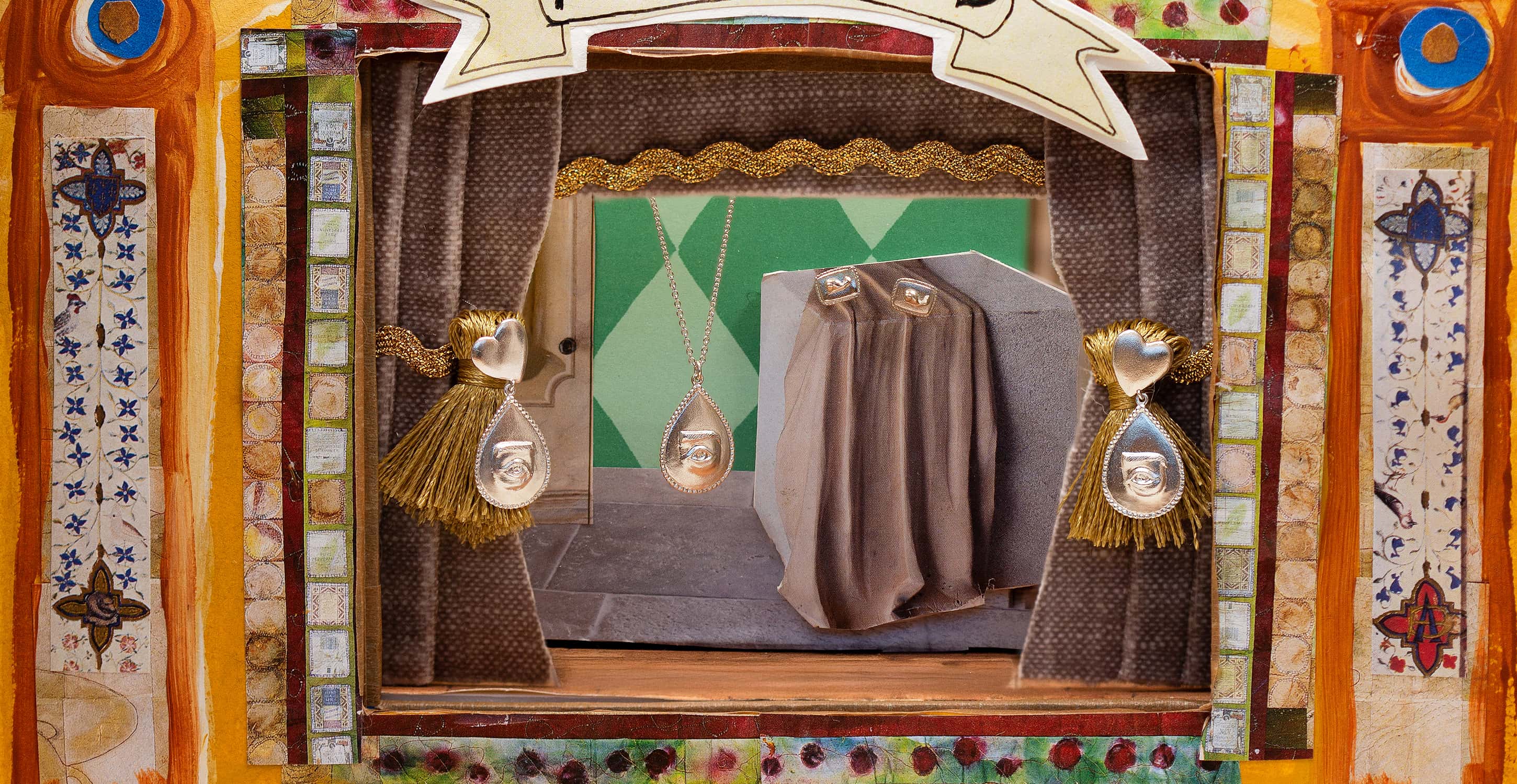 Handmade jewellery displayed on a miniature stage set, featuring gold-tone tassel earrings and teardrop pendants framed by patterned curtains and decorative panels.