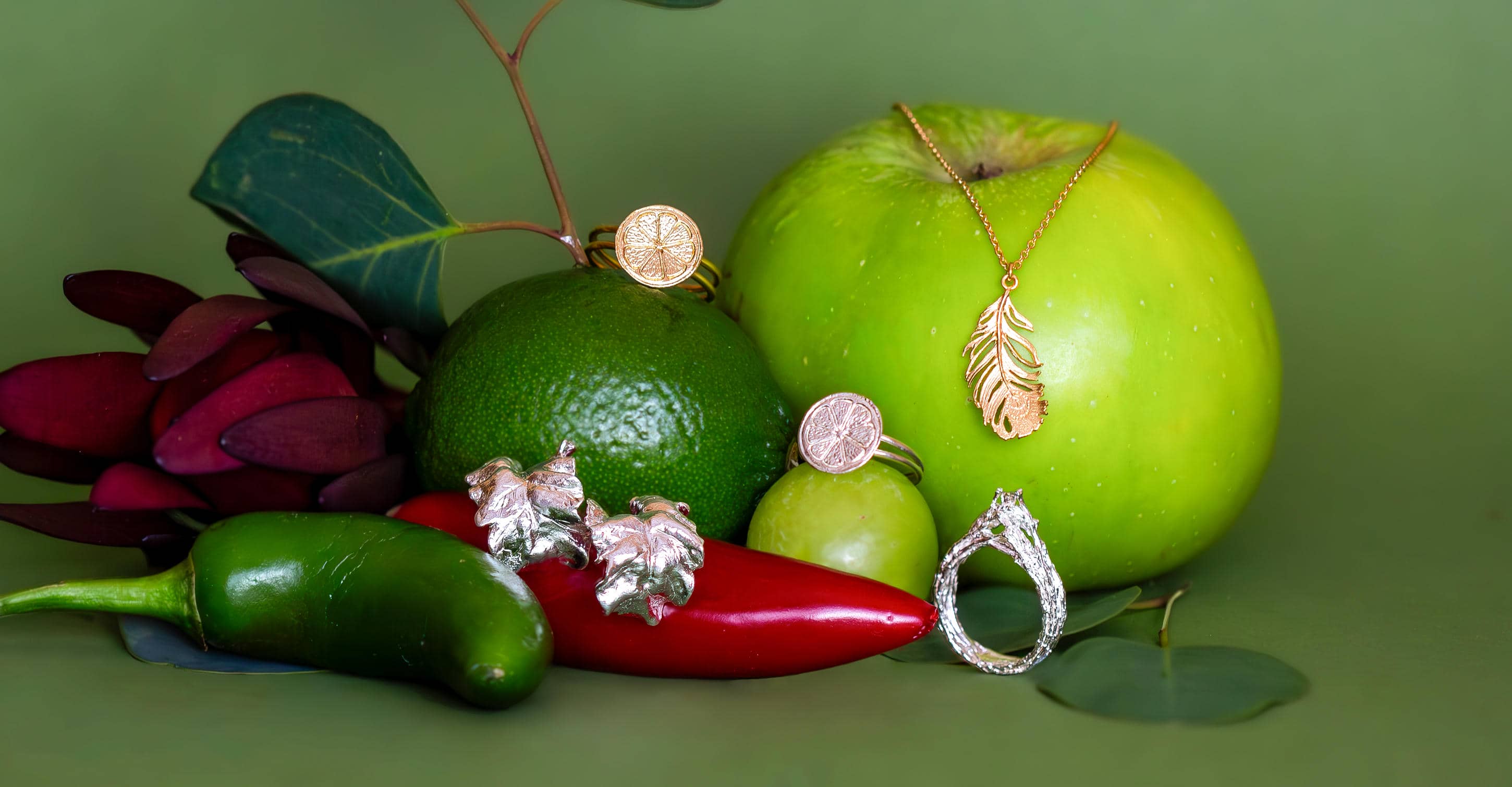 A still life image with green background and assorted mostly green fruits, with a few red flourishes. Jewellery is displayed on the fruits, including a feather necklace and chard earrings.